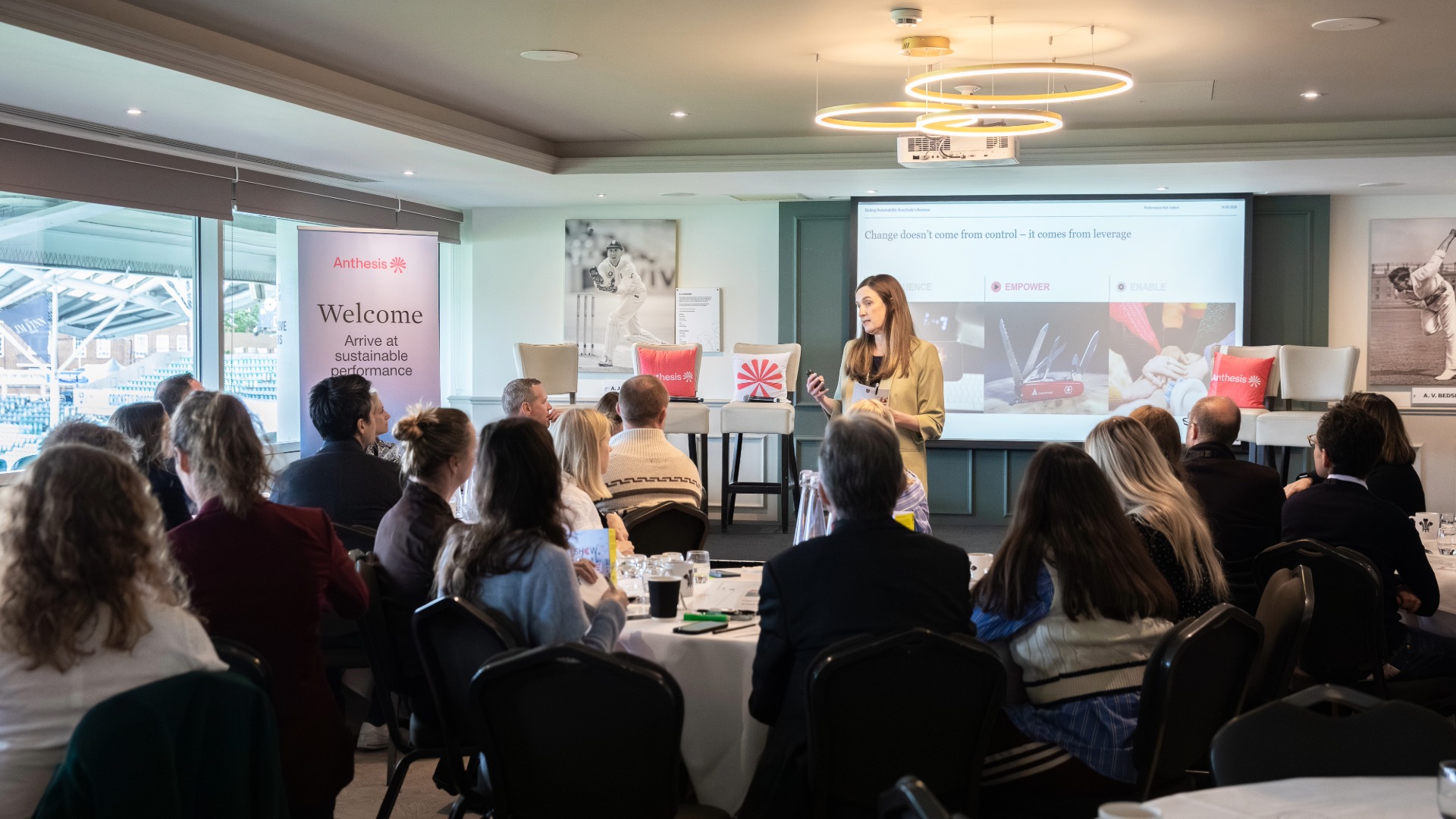 Anthesis Activator Summit: Show Me the Performance A woman stood in front of a number of tables of people, talking