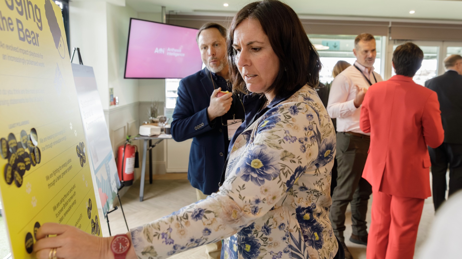 Anthesis Activator Summit: Show Me the Performance A woman sticking a sticker onto a yellow A1 placard with people in the background